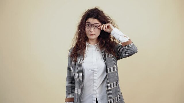 The young woman with curly sleepy hair, helmet and has a state of drowsiness. Young hipster in gray jacket and white shirt, with glasses posing isolated on beige background in the studio.