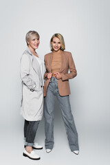 full length view of mother and daughter in stylish outfit smiling at camera on grey