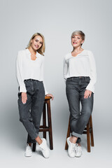 Obraz premium full length view of smiling mother and daughter in trendy clothes posing near high stools on grey