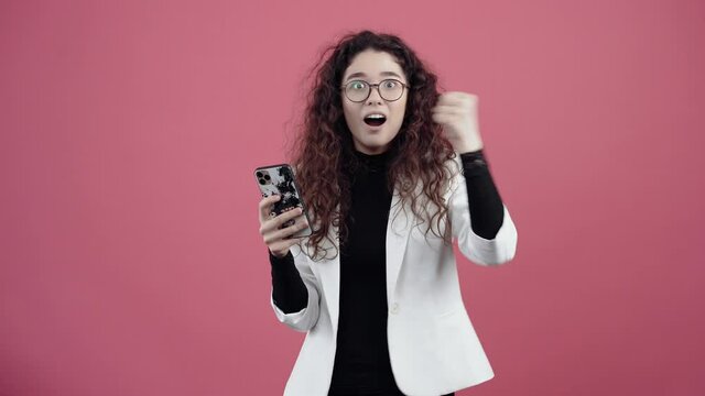 Happy young woman with curly hair, holds her mobile phone in her hand, and looks at the spray, smiling. Young hipster in white jacket and black shirt,