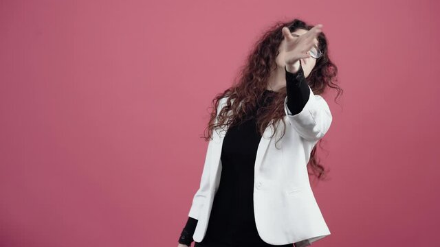 Angry young woman with curly hair, she looks with her hand to push away because she can't see. Young hipster in white jacket and black shirt, with glasses posing isolated on pink background in the