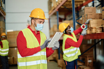Caucasian adult warehouse worker checks delivery order on a tablet while wearing a safety mask -...