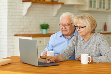 Happy aged couple sitting with hot drinks and watching movie or comminicating online on laptop in kitchen at home together. Elderly people active happy lifestyle, online communication concept