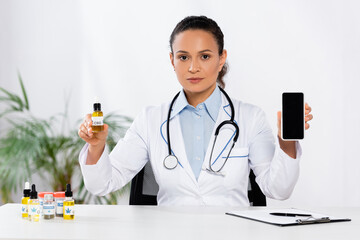 african american doctor holding bottle with cbd and smartphone with blank screen