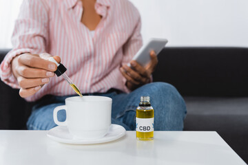 cropped view of african american woman holding smartphone and pipette with cbd liquid near cup of tea