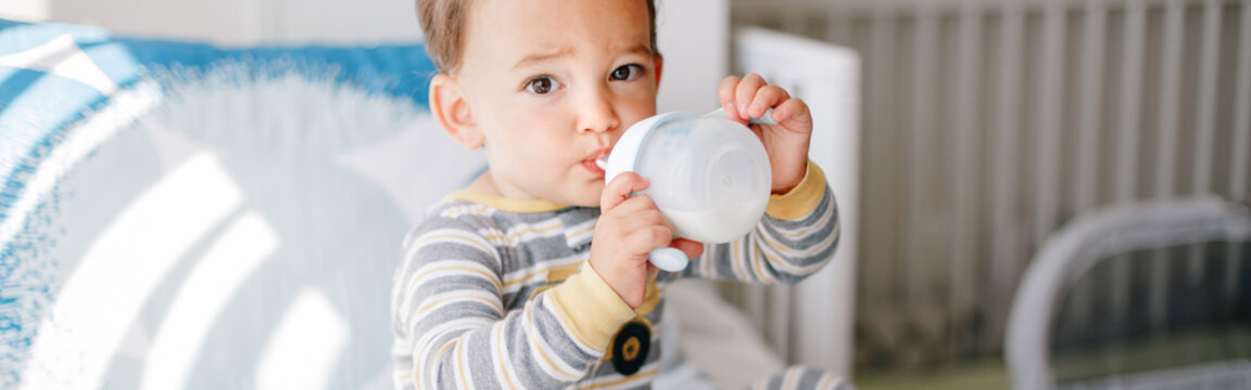  Kid boy sitting on bed drinking milk from kids bottle. Healthy eating drinking for little children. Supplementary food for growing babies. Candid real authentic moment. Web banner header.