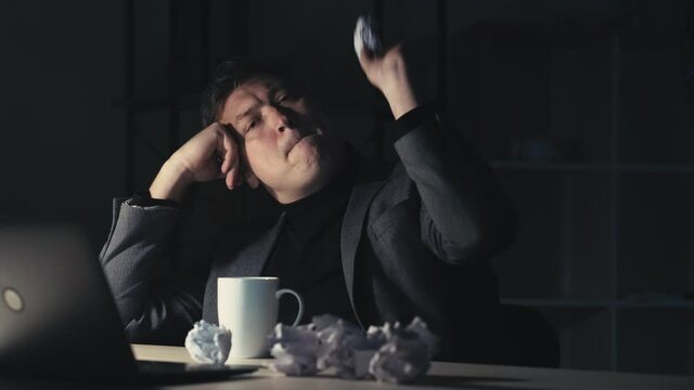 Procrastination crisis. Night work. Project postponement. Unproductive time. Relaxed lazy bored irresponsible male employee messing around throwing creased paper balls at dark home office workplace.