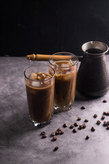 A coffee maker lined up in a row and two tall glasses with cold coffee with milk and ice on a dark background and scattered grains of natural coffee. Copy space.