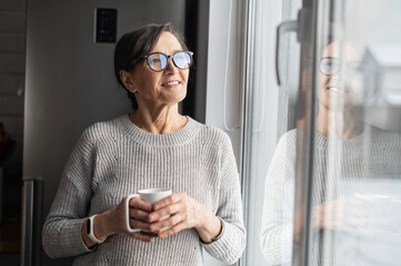 Positive and healthy modern senior woman holding mug with hot drinks, standing at kitchen home, looking through window, charming elderly female dreaming with cup of morning coffee