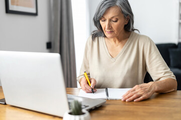 Focused retired woman is learning online, using a laptop for watching online classes, webinar, senior female takes a notes sitting in the kitchen at home