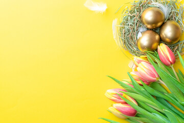 Obraz premium Easter eggs gold. Golden colour egg in basket with spring tulips, white feathers on pastel yellow background in Happy Easter decoration. Congratulatory easter design. Flat lay, top view.