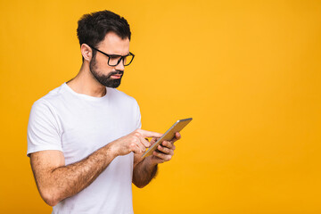 Happy young bearded man in casual standing and using tablet isolated over yellow background.