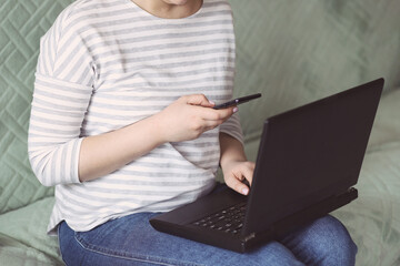 A young woman in a striped sweatshirt and blue jeans sits on the couch at home with a mobile phone in her hands and a laptop and works remotely online. Concept: freelancer. Selective focus.