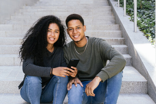 Lovely multiracial couple together sitting on building staircase, smiling biracial woman and cheerful African American boy hold smart phone, look at webcam. Couple are surfing the web on the phone.