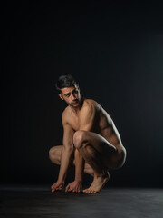 Portrait of a naked male model posing squatted on black studio background