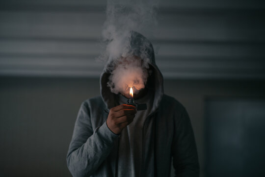 Unrecognizable male in hooded jacket smoking cigarette while standing in dark room with burning lighter in hand