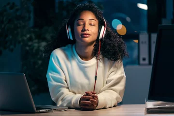 Tuinposter Confident african woman listening to music with earphones while relaxing of work in the office at home. © nenetus
