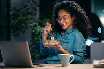 Beautiful afro young business woman working with computer while eating pasta sitting in the office.