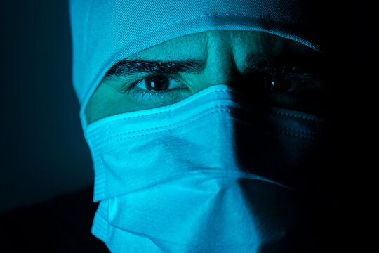 Closeup of male surgeon in medical mask looking at camera in dark room with blue neon light