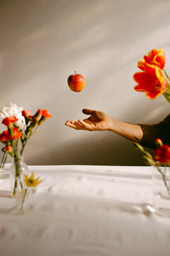 Crop anonymous person tossing ripe apple in air above table with tulips and fresh carnations