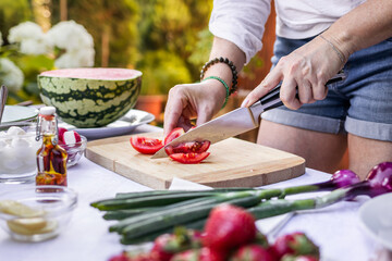 Woman is chopping red tomato on cutting board for vegetable salad. Preparing mediterranean dish for garden party