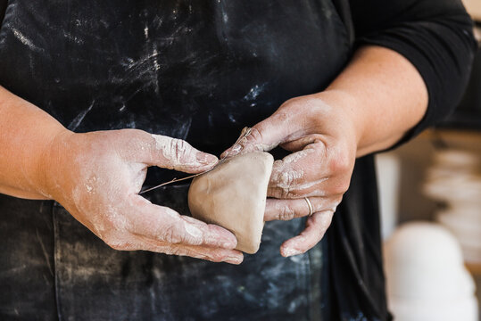 Crop Anonymous Master In Dirty Apron Standing In Workshop And Shaping Piece Of Clay Marking It With Flower In Hands