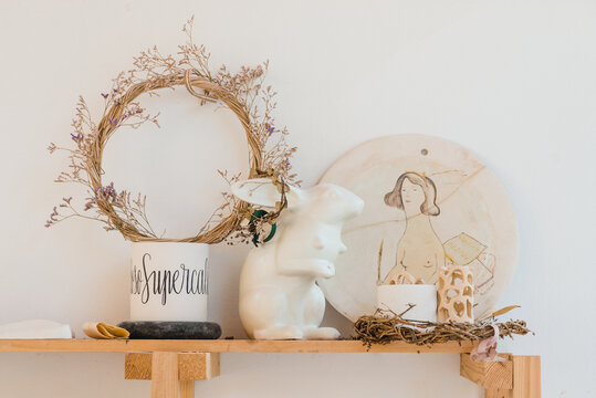 Wreath with dry plants near bunny statuette and hand painted chopping board near cup and decorative objects on wooden shelf near white wall