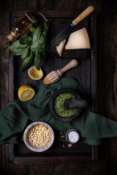 Top view composition with ingredients for traditional pesto sauce including Parmesan cheese and basil leaves and lemon and pine nuts arranged on dark table