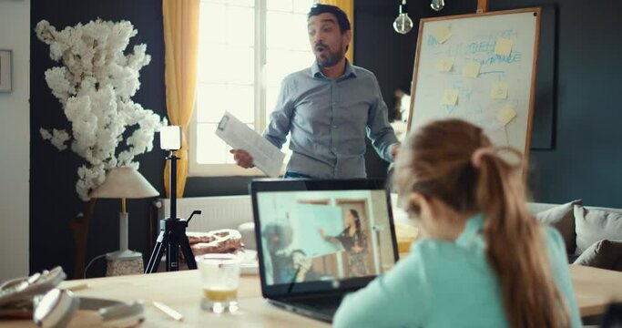 Father And Daughter Work From Home Online At The Same Time. The Girl Looks At The Teacher On Her Laptop And Learns School From Home. A Man Broadcasts A Live Video Conference Using A Telephone