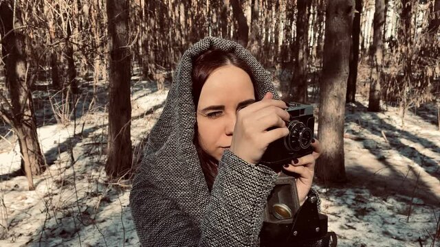 Young woman in gray coat photographing on old photo camera in forest. Pretty female taking photos with old camera in early spring.