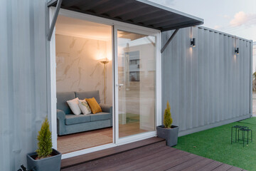 Contemporary gray container house with comfy sofa behind glass door located on grassy yard
