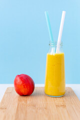 Fototapeta premium Smoothie or cocktail of nectarine, almond or oat milk and turmeric in a glass jar with two cocktail tubes. Juicy ripe nectarine on a cutting board on a blue background. Copy space, vertical.