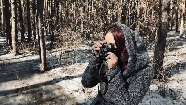 Young woman in gray coat photographing on old photo camera in forest. Pretty female taking photos with old camera in early spring.