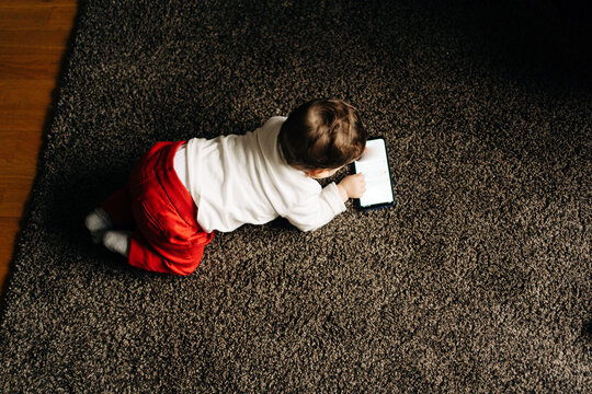 From above content little baby boy lying on fluffy carpet and watching funny video on mobile phone in light living room