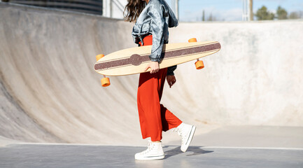 Side view of cropped unrecognizable happy young female in stylish apparel walking with longboard on concrete skate park