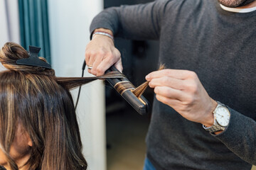Crop unrecognizable male stylist in casual clothes using hair straightener while doing curls for female client in beauty salon