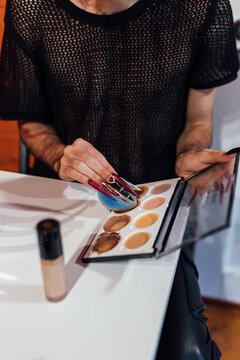 Crop anonymous transsexual man with sponge and cream foundation palette preparing for makeup at table indoors