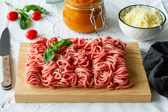 From above of fresh raw mixed pork and beef minced meat on wooden cutting board with green basil leaves placed on table during cooking process near homemade tomato sauce