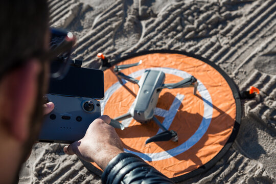 From above of unrecognizable male with remote controller standing on beach with drone placed on landing pad