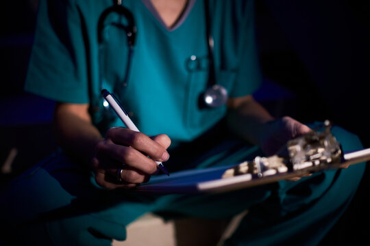 Cropped unrecognizable female medic in uniform reading documents on clipboard while analyzing medical report in dark room
