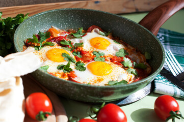 Traditional dish of israeli cuisine Shakshuka. Fried eggs with tomatoes, peppers, onions, cilantro and pita in a pan on light green background. Top view