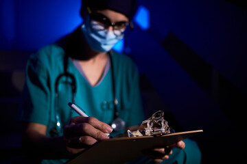 Unrecognizable female medic in mask and uniform reading documents on clipboard while analyzing medical report in dark room