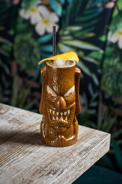 Brown sculptural tiki mug with alcohol drink decorated with straw and ice placed on table on blurred background