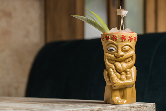 Polynesian tiki cup of cold alcohol beverage decorated with straw and green pineapple leaves placed against on wooden table