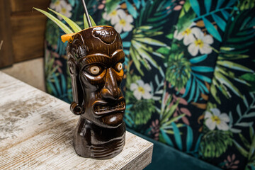 Brown sculptural tiki mug with alcohol drink decorated with straw and ice placed on wooden table