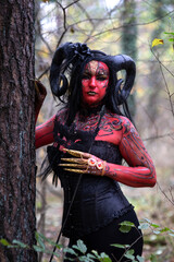 Naklejka premium Sexy Teufelin steht im Wald. Sie hat die Hörner auf dem Kopf, eine schwarze Corsage. sie ist mit der roten Farbe und der Muster angemalt.Sie hat lange, schwarze Haare. Nah Portrait.