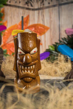 Brown sculptural tiki mug with alcohol drink decorated with straw and ice placed amidst dry grass against green plant with feather on blurred background