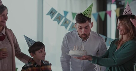 Cheerful bearded adult father blowing candles on birthday cake celebrating in family circle. Six caucasin family members gathering around to spend weekend.