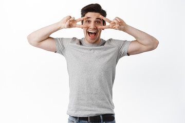 Excited happy man in t-shirt showing peace signs, v-sign kawaii over eyes and looking joyful, smiling, standing over white background