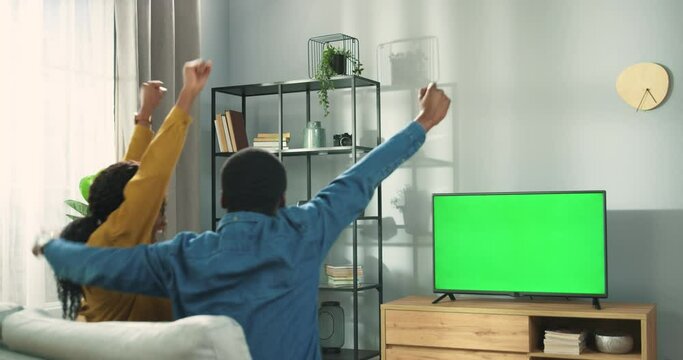 Rear of happy African american couple man and woman sitting on couch in living room watching at TV monitor with green screen in positive mood. Family cheering for fav team, watching TV, sport match